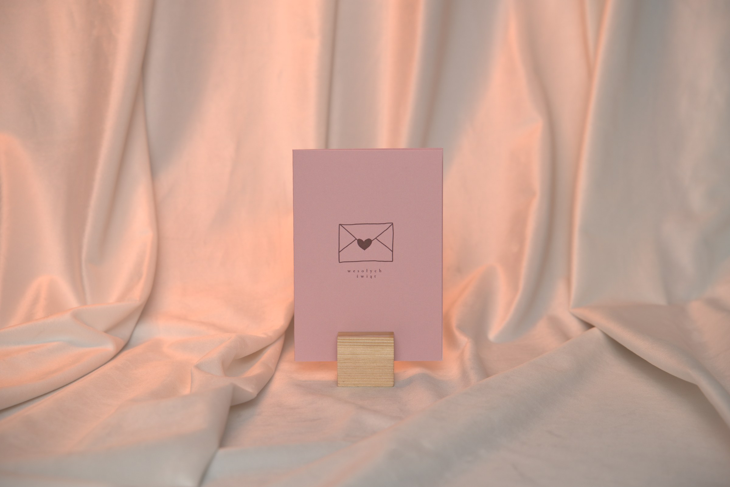Różowa kartka świąteczna z motywem koperty z sercem i napisem Wesołych świąt na drewnianym stojaku na kremowym atłasie