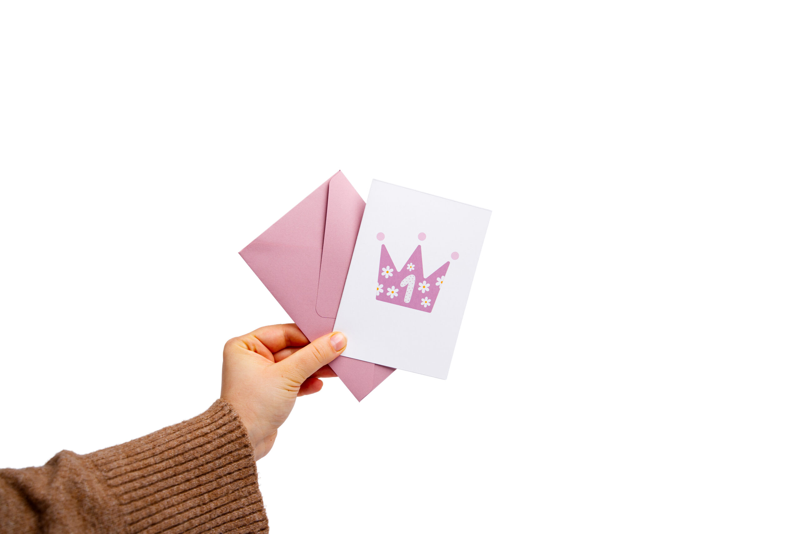 Biała kartka na roczek z różową koroną i różową kopertą trzymana w dłoni