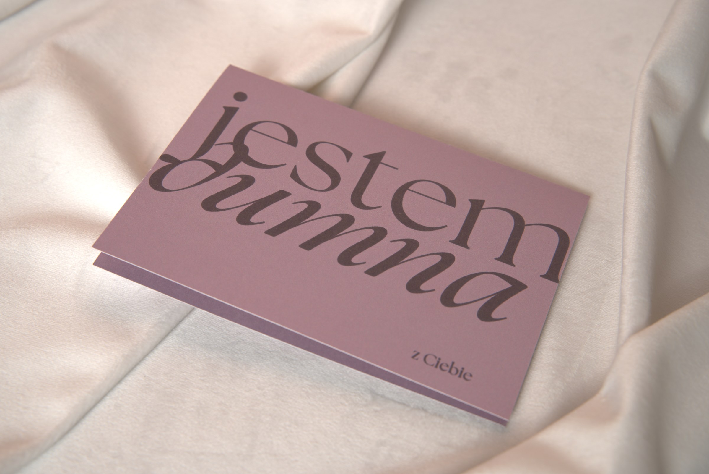 Liliowa kartka gratulacyjna z kaligraficznym napisem Jestem dumna z Ciebie ułożona ukośnie na kremowym atłasie – flat lay